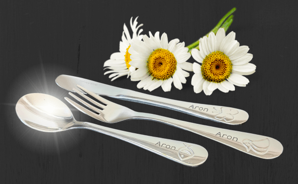 Personlig gravert barnebestikk med navnet Aron, dekorert med symboler, lagt ved tre prestekrager på sort bakgrunn.